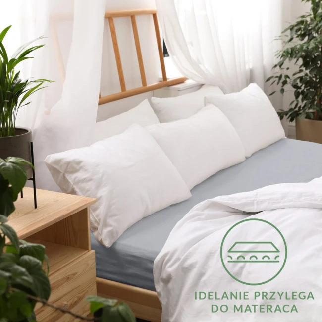 Prześcieradło jersey z gumką 70x140 w kolorze popiel na łóżku, z lewej strony widoczne białe poduszki. Produkt leży na materacu, z delikatnym grubym materiałem, który idealnie przylega. W tle rośliny i drewniane elementy, logo i tekst w dolnym rogu.