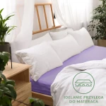 Prześcieradło jersey z gumką 80x160 w kolorze jasnym fiolecie, rozłożone na łóżku. Widoczne białe poduszki i miękka narzuta. Tło naturalne z drewnianymi elementami oraz roślinnością, z logo „Idealnie przylega do materaca” w rogu.