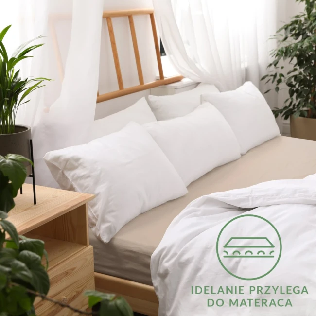 Beżowe prześcieradło jersey z gumką w rozmiarze 80x160, rozłożone na łóżku, dobrze przylegające do materaca. W tle widoczne białe poduszki i drewniana rama łóżka. Delikatne tło z roślinnością i zasłonami.