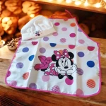 Fartuszek dziecięcy z czapką szefa, biały z kolorowymi kropkami, w centralnej części widoczny wzór postaci Minnie w różowej sukience. Czapka szefa w białym kolorze z haftem. Fartuszek zawieszony, widok frontalny, z długimi paskami do związania.