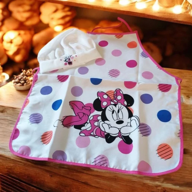 Fartuszek dziecięcy z czapką szefa, biały z kolorowymi kropkami, w centralnej części widoczny wzór postaci Minnie w różowej sukience. Czapka szefa w białym kolorze z haftem. Fartuszek zawieszony, widok frontalny, z długimi paskami do związania.