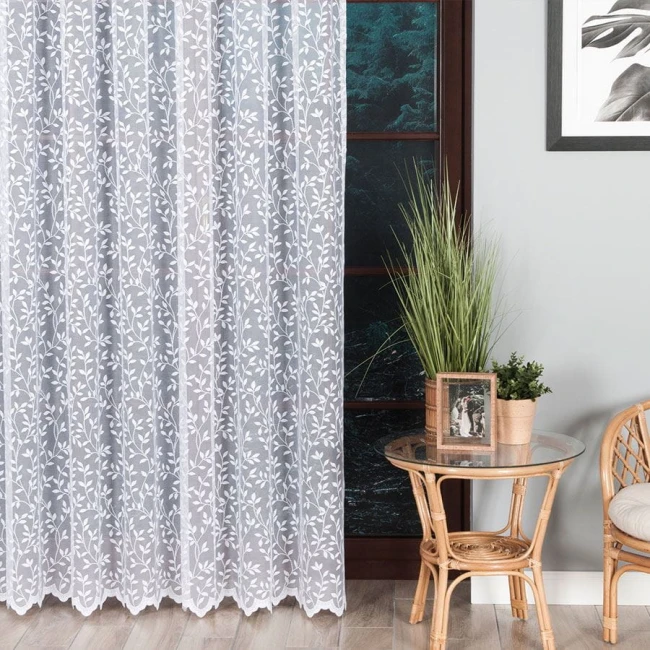 Firana żakardowa Zuzanna w kolorze białym o wysokości 250 cm, z delikatnym wzorem liści. Produkt wiszący, zbliżenie na część firany, w aranżacji wnętrza z rattanowym stolikiem, roślinnością w doniczce i zdobioną ramką na zdjęcia.