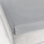 Prześcieradło żakardowe bez gumki w kolorze popielatym, o wymiarach 175x220 cm, leżące na łóżku. Detal przedstawia delikatny wzór, teksturę i fakturę materiału. Tło jasne, prostokątne, prezentujące estetykę aranżacji sypialnianej.