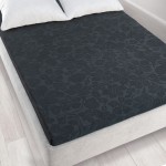 Prześcieradło żakardowe bez gumki w kolorze grafitowym o wymiarach 150x205 cm, leżące na łóżku. Wzór w formie kwiatów, tekstura dzianiny, z widocznymi poduszkami w tle. Tło w neutralnym kolorze, podkreślające wygląd produktu.