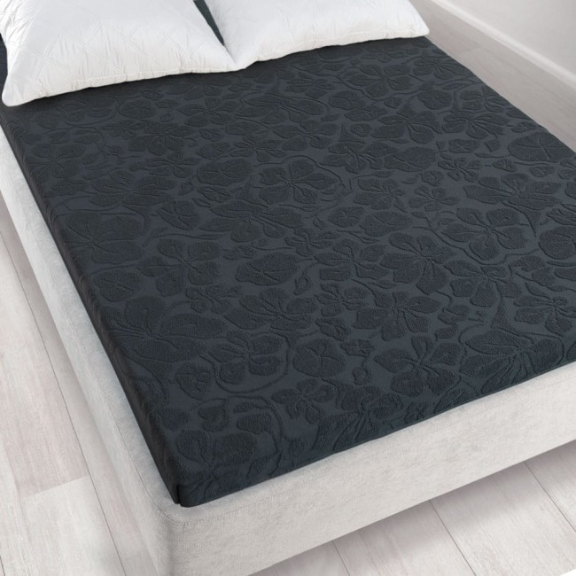 Prześcieradło żakardowe bez gumki w kolorze grafitowym o wymiarach 150x205 cm, leżące na łóżku. Wzór w formie kwiatów, tekstura dzianiny, z widocznymi poduszkami w tle. Tło w neutralnym kolorze, podkreślające wygląd produktu.