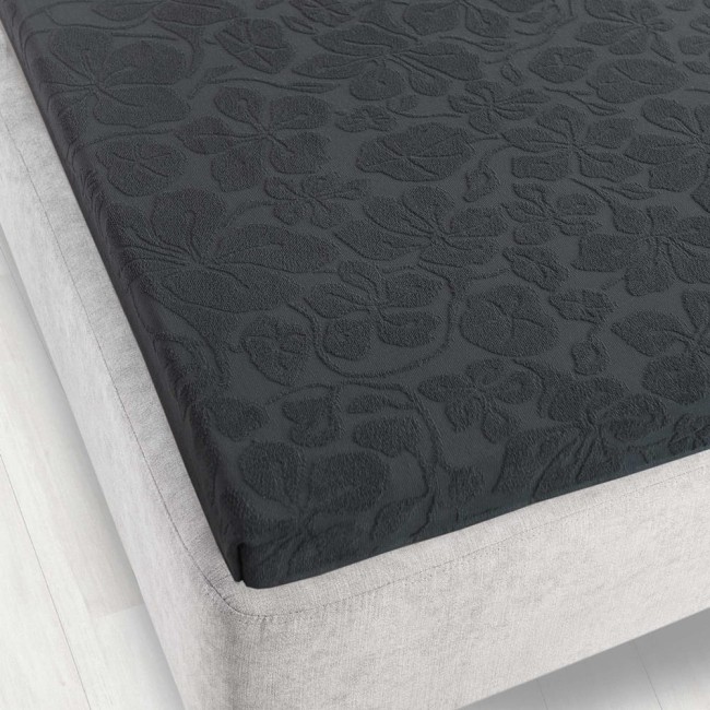 Prześcieradło żakardowe bez gumki 150x205 w kolorze grafitowym. Widok z góry, na rozłożonym materacu. Wzór kwiatowy o delikatnej fakturze, matowy materiał, dobrze dopasowane do krawędzi. Tło w jasnym odcieniu dla lepszej widoczności produktu.